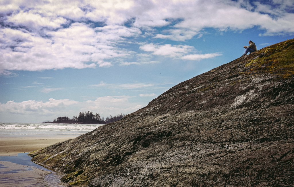 A man sits on the top of a rocky outcrop looking out to sea