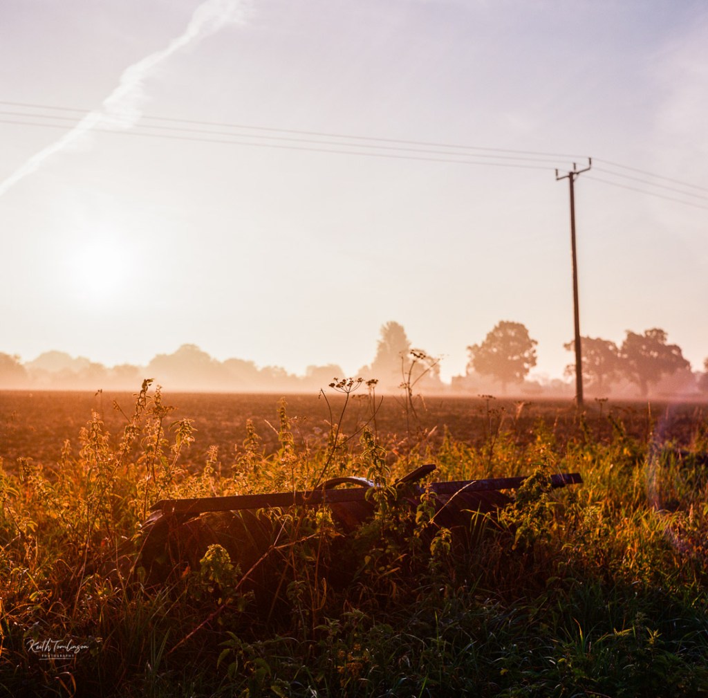 Sun rises over a ploughed field with a plough resting at the edges of the field
