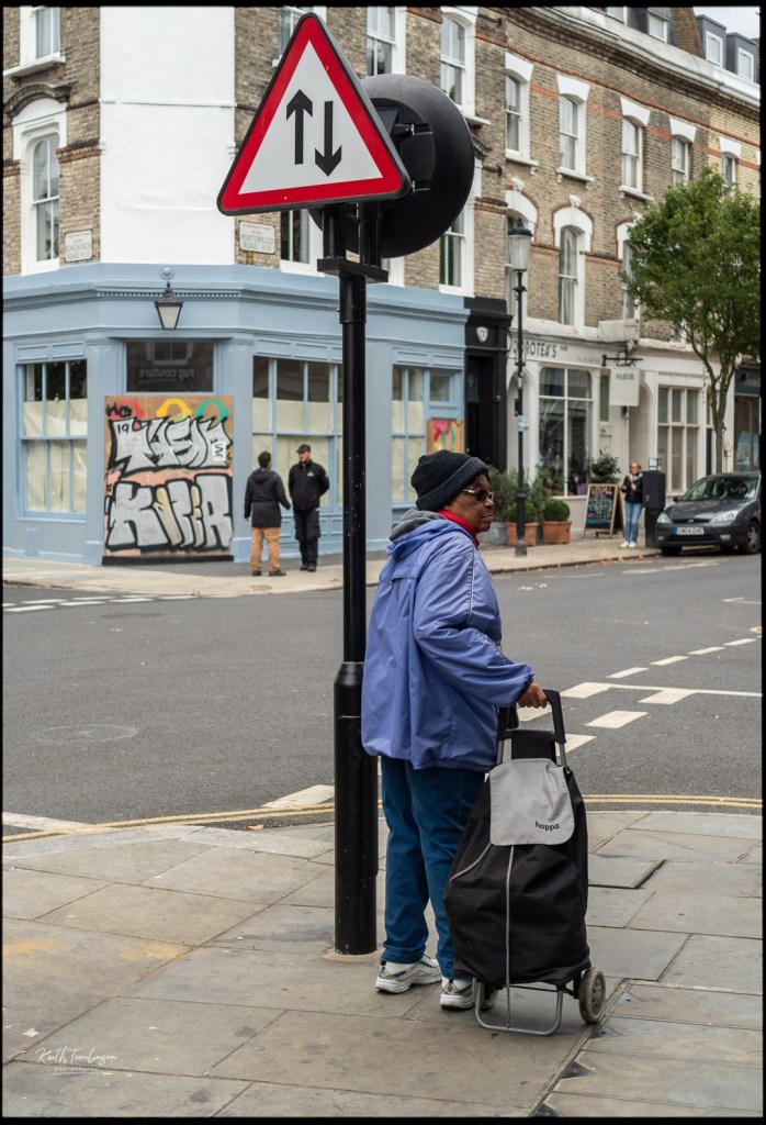 A  lady waits by the end of the road with her shopping trolley