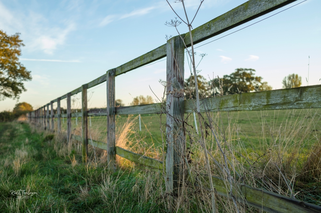 A wooden fence at the end of a field is seen close to the viewer and then disappears into the distance