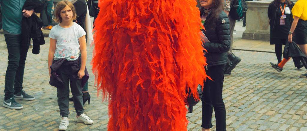A child looks quizzically at a man in high heeled boots and a large red feathered jacket