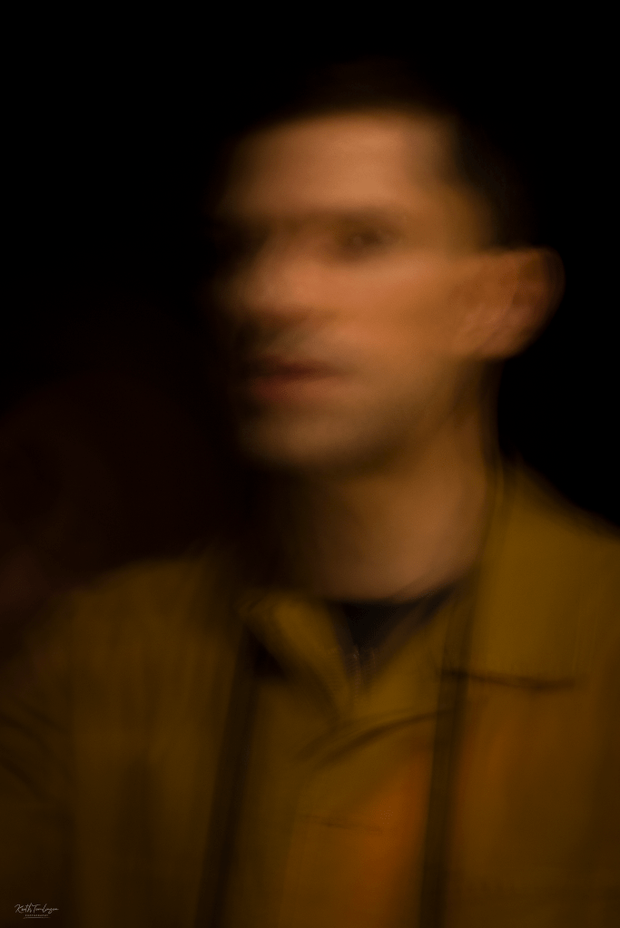 A blurred photo of a man with black hair in a dark room, staring at the photographer