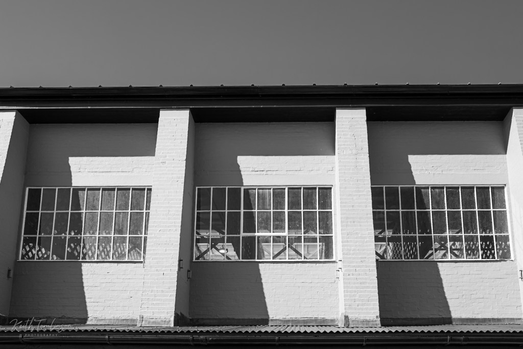 Black and white photograph of the exterior upper storey of a 1920s aircraft hangar at IWM Duxford, showing three large multi-pane industrial windows set into painted brick bays, with strong diagonal shadows cast by the afternoon sun.