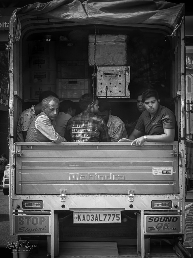 Black and white street photograph of men riding in the back of a Mahindra truck in Bengaluru, India